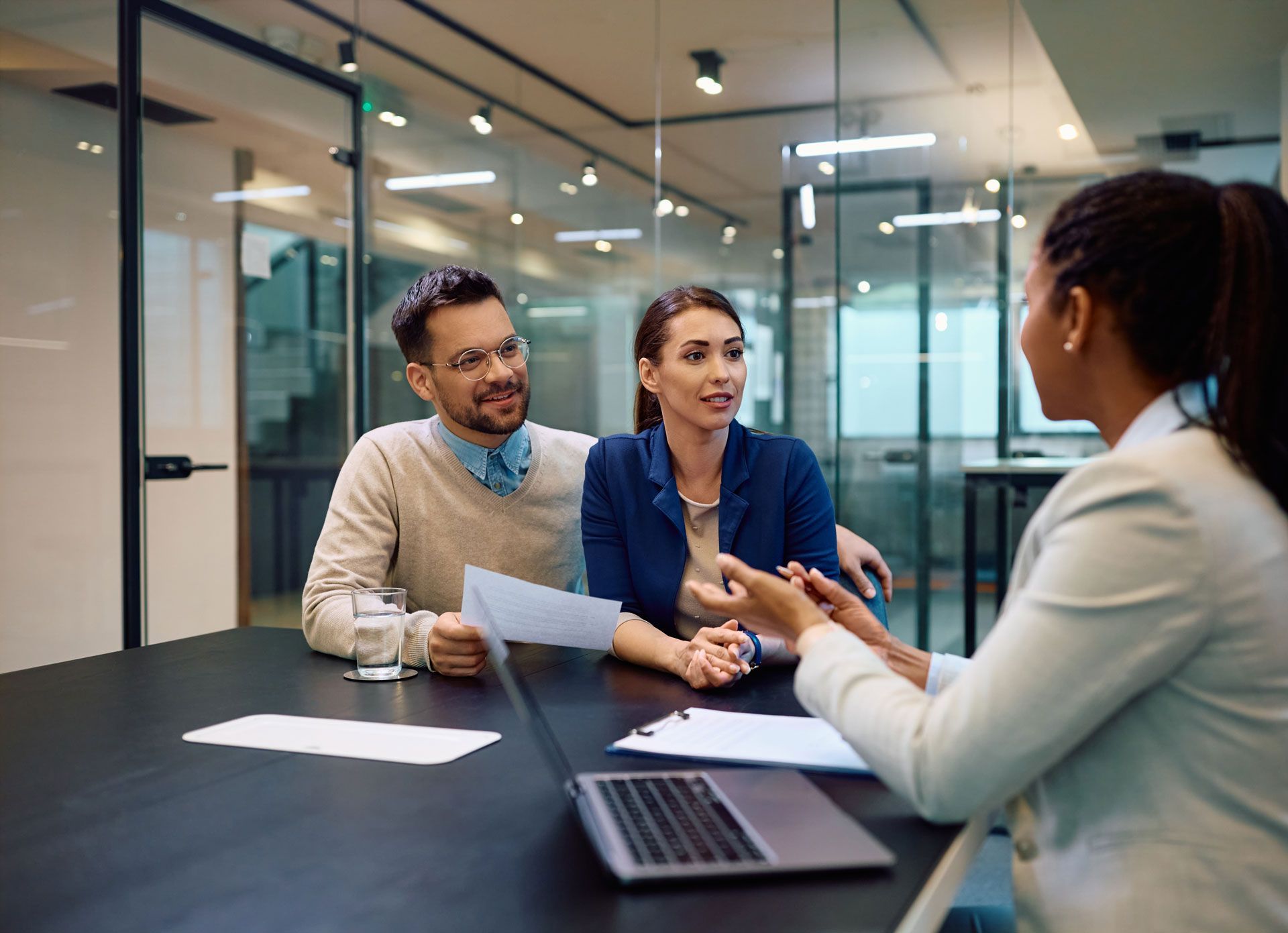 Couple at a table with a professional, reviewing documents. Inside a modern office, glass walls.