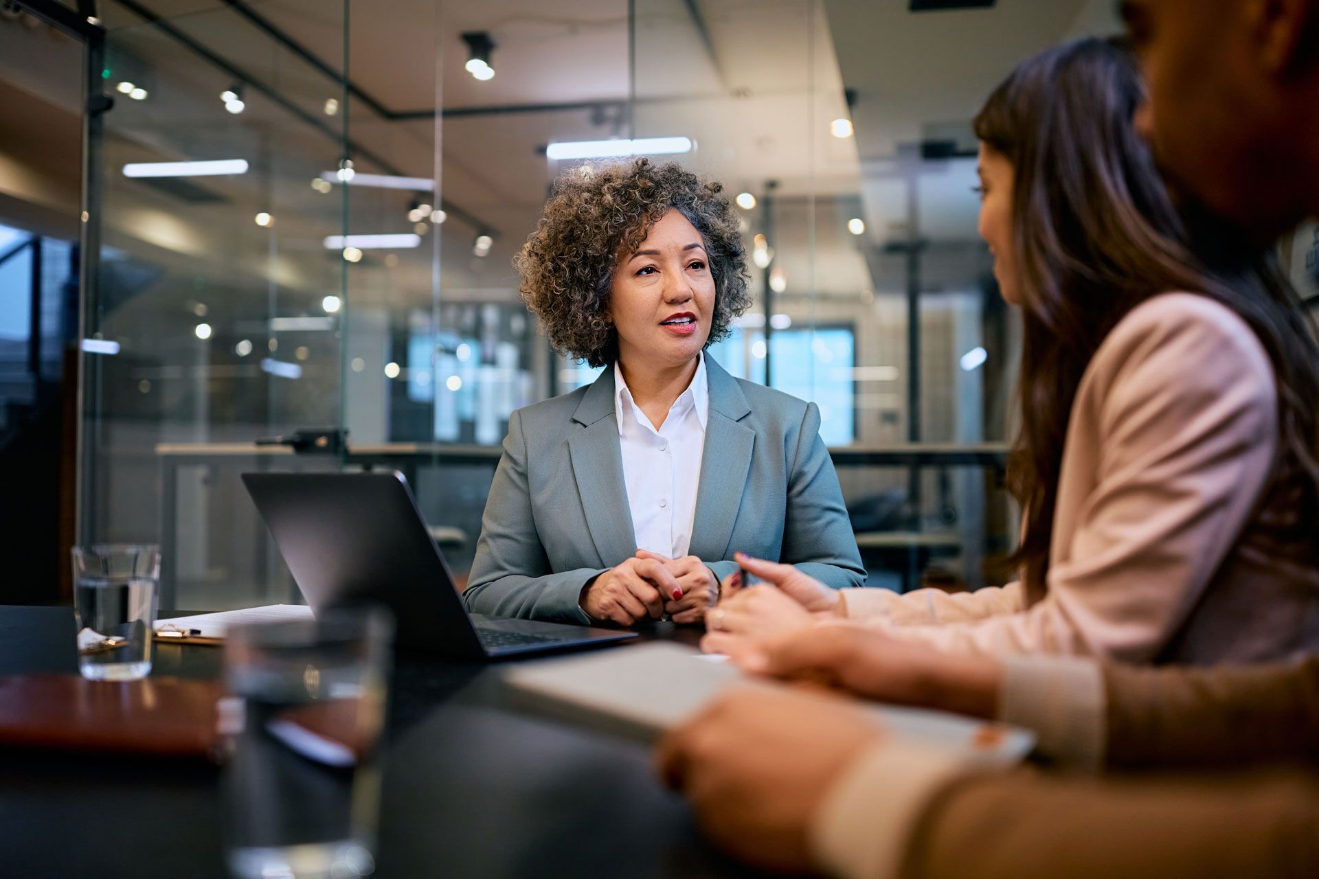 Woman in blazer talks to two people at a table, laptop in front, modern office setting.