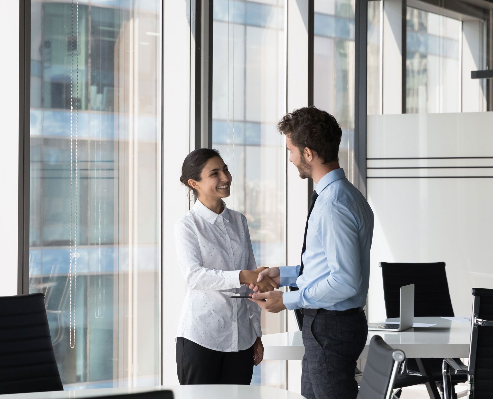 Two people in business attire shaking hands by a window in an office setting.