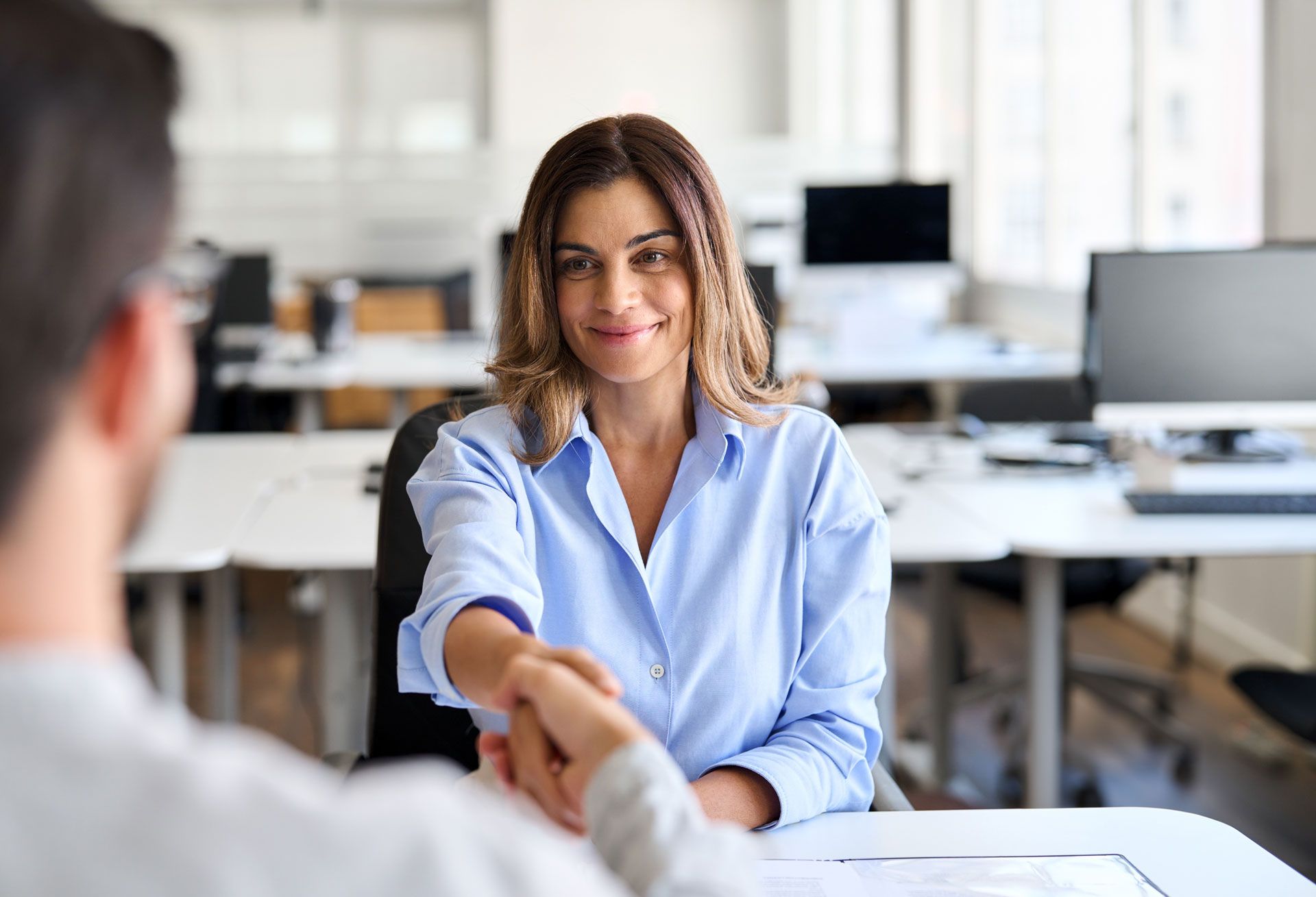 Woman in a light blue shirt shaking hands with a person in an office setting, smiling.