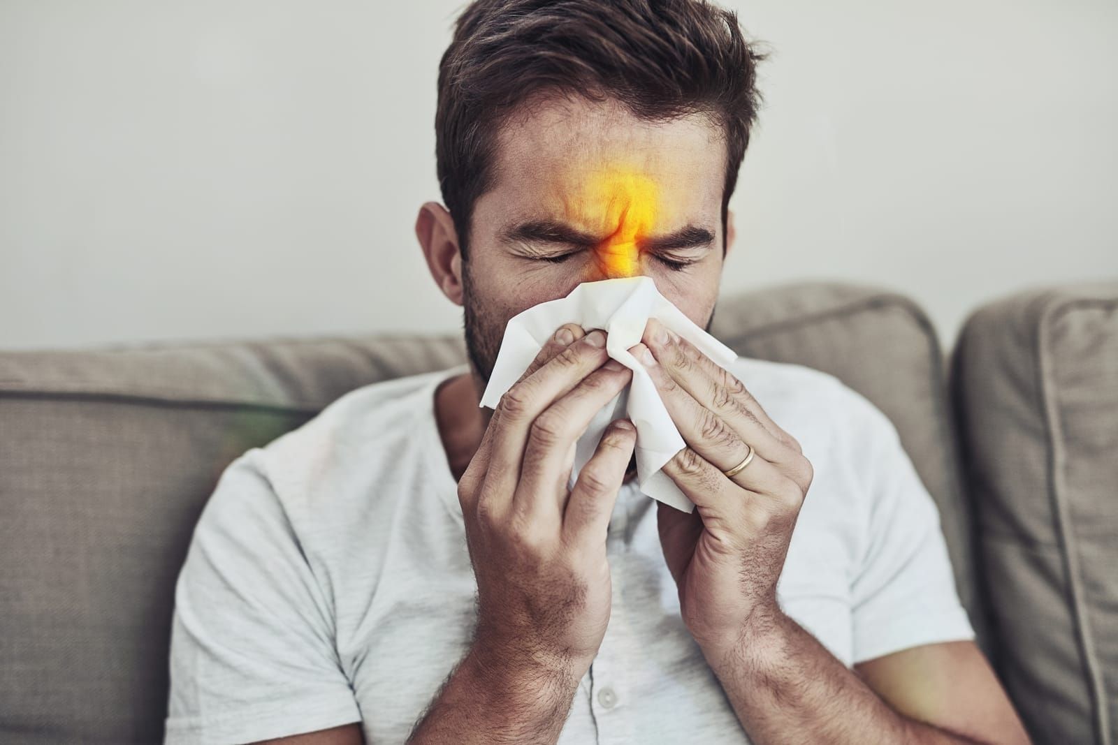 A man is blowing his nose into a napkin while sitting on a couch.