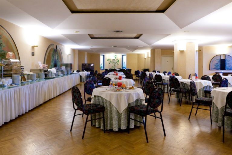Sala da pranzo con tavoli rotondi ricoperti da tovaglie bianche, sedie e un lungo tavolo da buffet addossato a una parete.