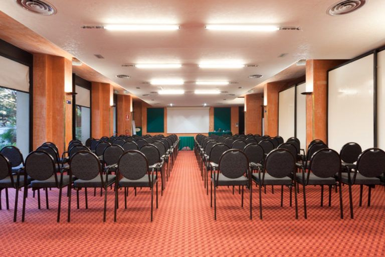 Una sala conferenze d'albergo con file di sedie nere disposte di fronte a uno schermo per presentazioni, illuminate da faretti a soffitto e poggiate su un tappeto rosso.