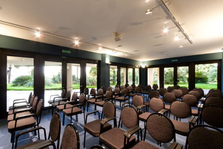 Una sala conferenze vuota con file di sedie scure imbottite di fronte a porte a vetri che si affacciano su un giardino.