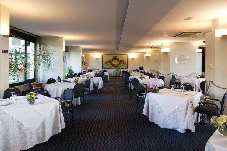 Una sala da pranzo spaziosa e ben illuminata, con tavoli apparecchiati con tovaglie bianche, sedie scure e un tappeto a motivi geometrici.