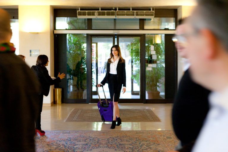 Una persona in abito da lavoro trascina una valigia viola attraverso l'ingresso della hall di un hotel, incorniciata da alcune persone in primo piano.