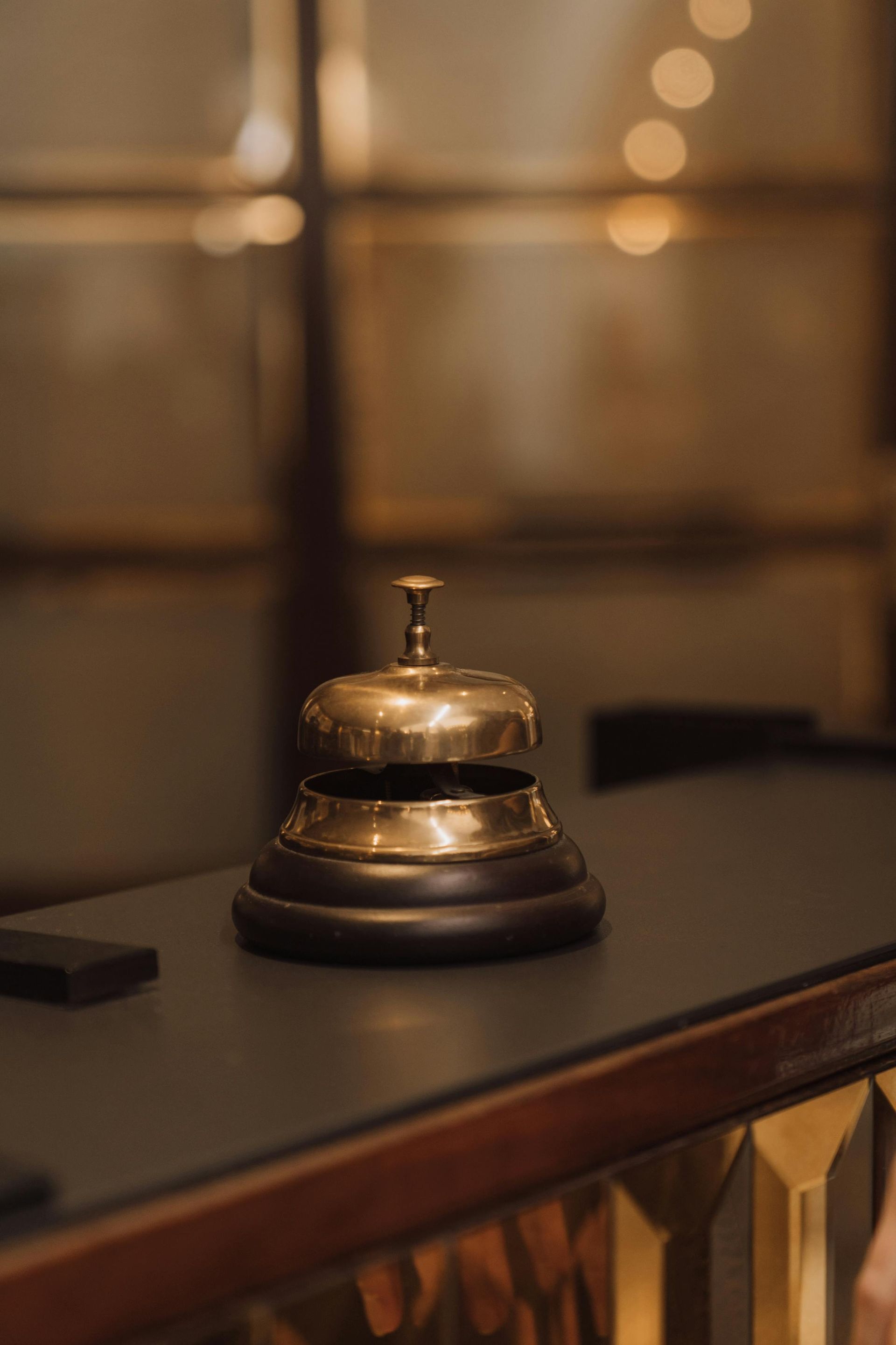 Su un bancone scuro della reception, in uno sfondo leggermente sfocato e illuminato da una luce soffusa, si trova un campanello di servizio in ottone lucido.