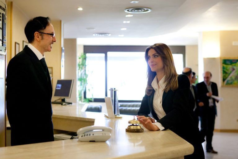 Due professionisti in abiti da lavoro sono in piedi alla reception di un hotel e conversano tra loro.