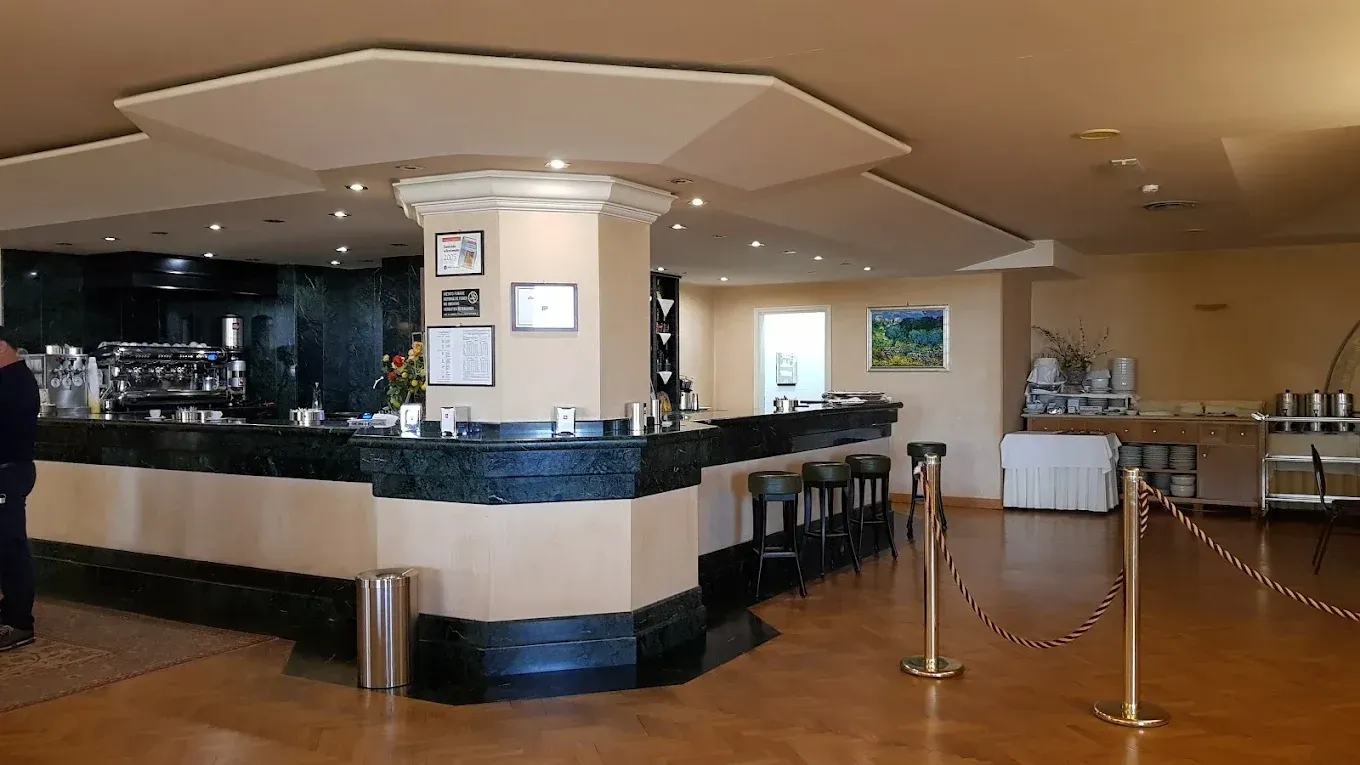 Il bar della hall di un hotel presenta un bancone beige con finiture nere, sgabelli e una corda di sostegno in un ambiente caldo e illuminato.