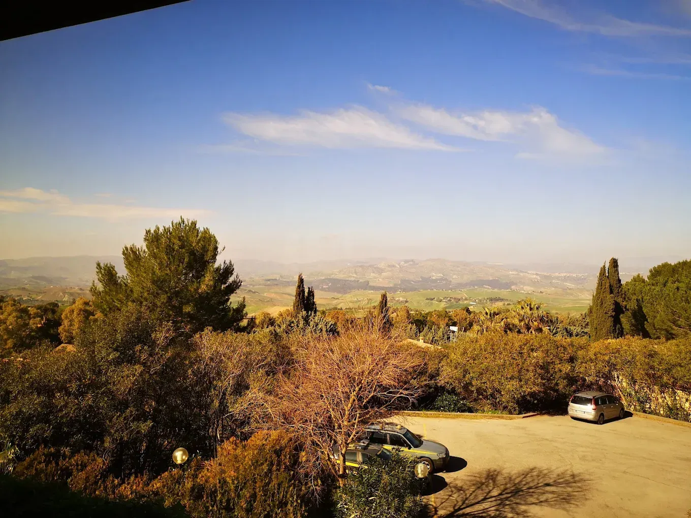 Una veduta panoramica di un paesaggio collinare e ondulato sotto un cielo azzurro, con in primo piano un parcheggio contenente due auto.