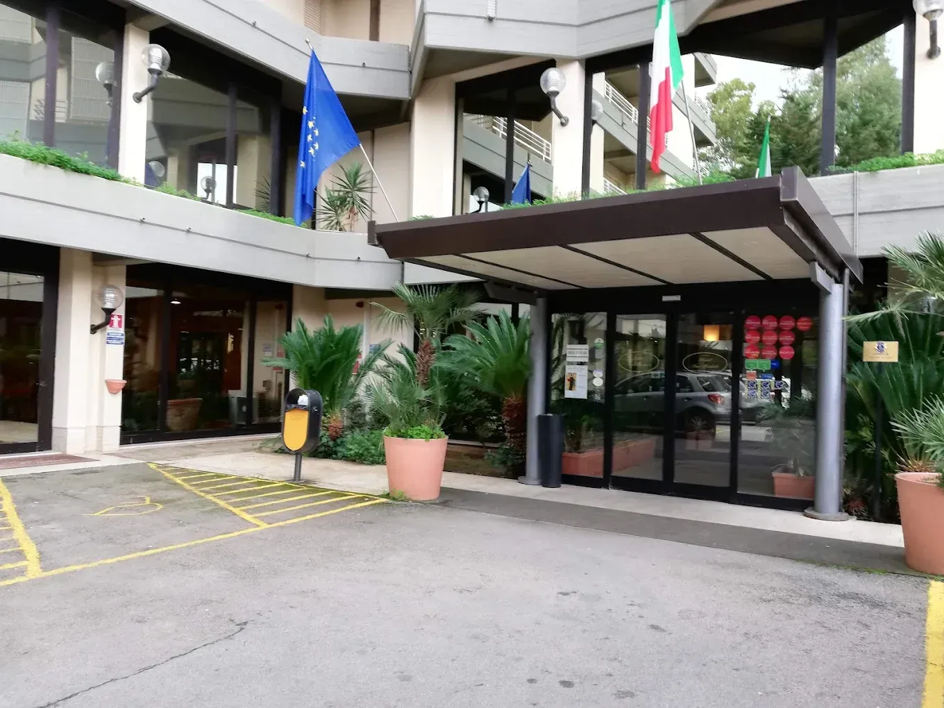 Ingresso di un moderno edificio per uffici con porte a vetri, piante in vaso e bandiere dell'Unione Europea e dell'Italia.