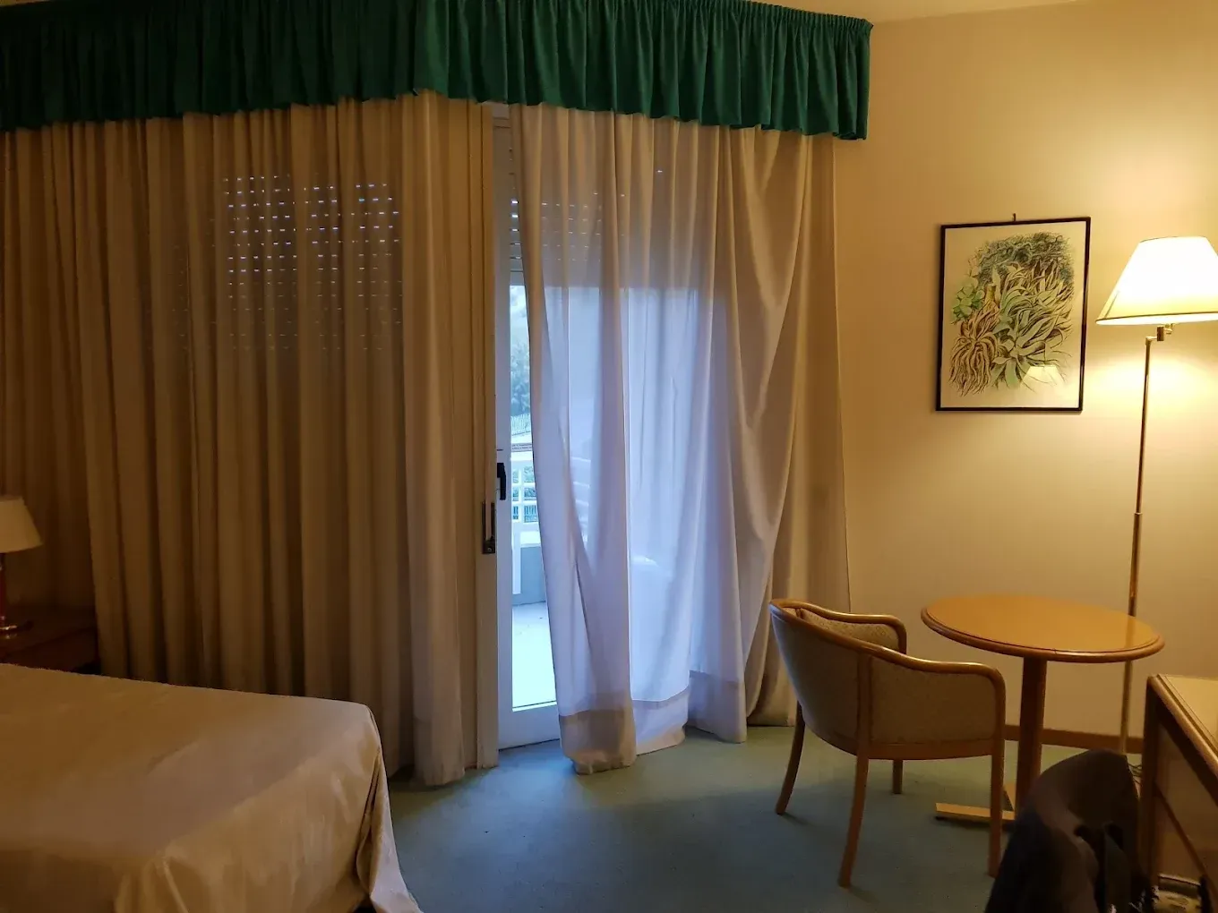Una camera d'albergo con un letto, un tavolino rotondo con una sedia, una lampada da terra e una grande finestra coperta da tende.
