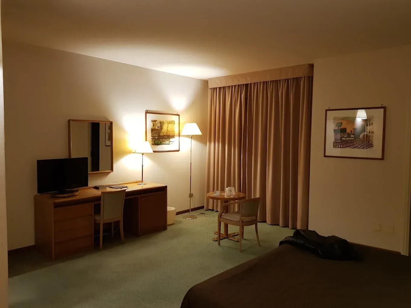 Una camera d'albergo con una scrivania in legno, televisione, una lampada, un tavolino rotondo con due sedie e un letto con lenzuola marroni.