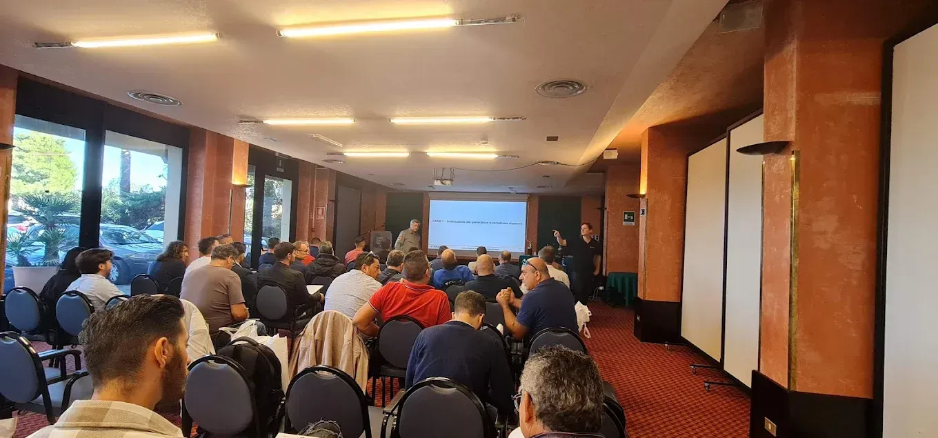 I partecipanti sono seduti in una sala conferenze con tappeto rosso e luci calde, di fronte al relatore proiettato su uno schermo.