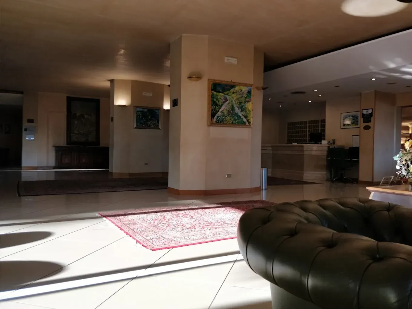 Una hall con un divano trapuntato in pelle scura in primo piano, un tappeto rosso su un pavimento chiaro e un banco della reception.