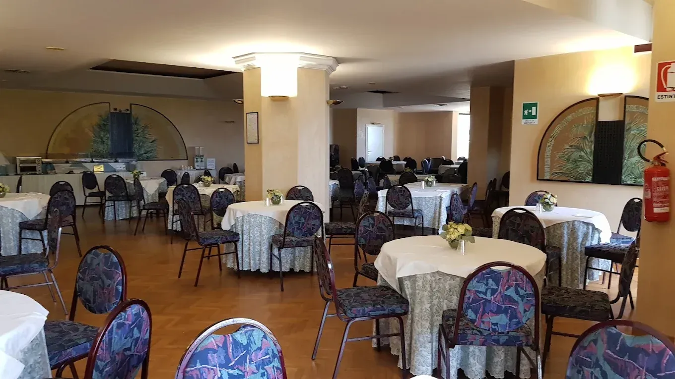 La sala da pranzo di un ristorante, con diversi tavoli rotondi ricoperti da tovaglie bianche e sedie decorate su un pavimento piastrellato.