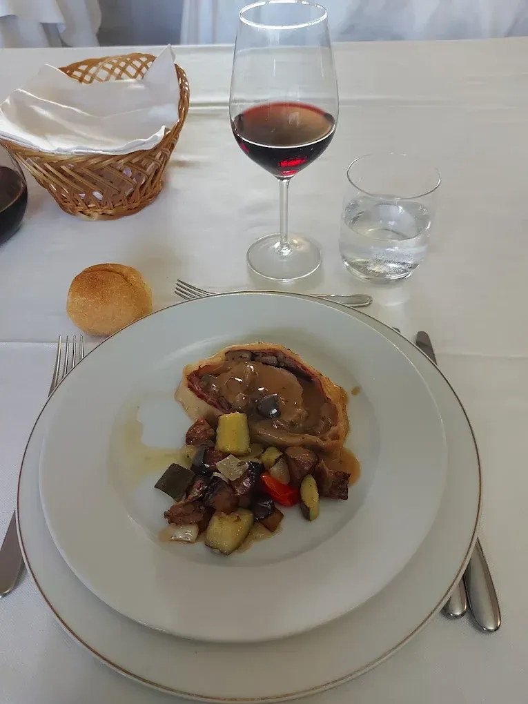 Su un tavolo, un piatto con verdure arrosto e carne in salsa, un panino, un bicchiere di vino rosso e un bicchiere d'acqua.