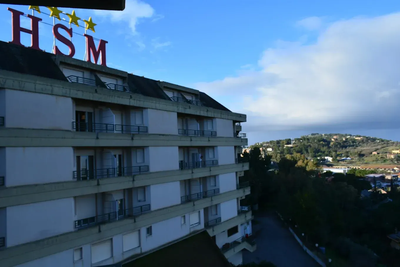 Un edificio a più piani dell'hotel HSM con balconi che si stagliano contro un cielo luminoso, affacciato su un paesaggio collinare.