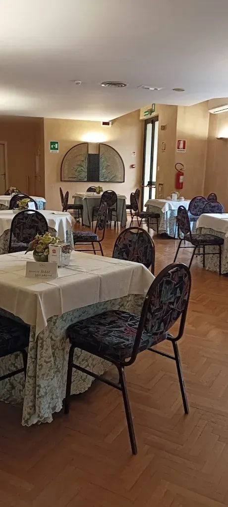 Una sala da pranzo con diversi tavoli quadrati ricoperti da tovaglie bianche, abbinati a sedie scure con motivi decorativi, su un pavimento in legno.