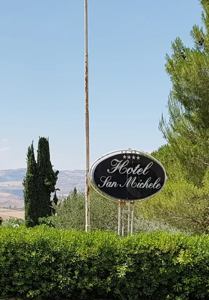Insegna ovale dell'Hotel San Michele con quattro stelle, montata su un palo sopra una siepe verde con uno sfondo di paesaggio collinare.
