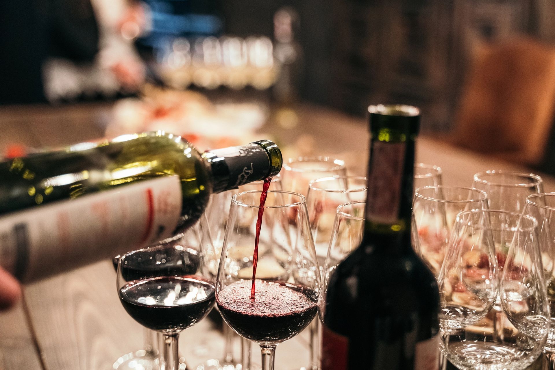 Vino rosso versato in un bicchiere durante una degustazione, con altri bicchieri e bottiglie visibili.