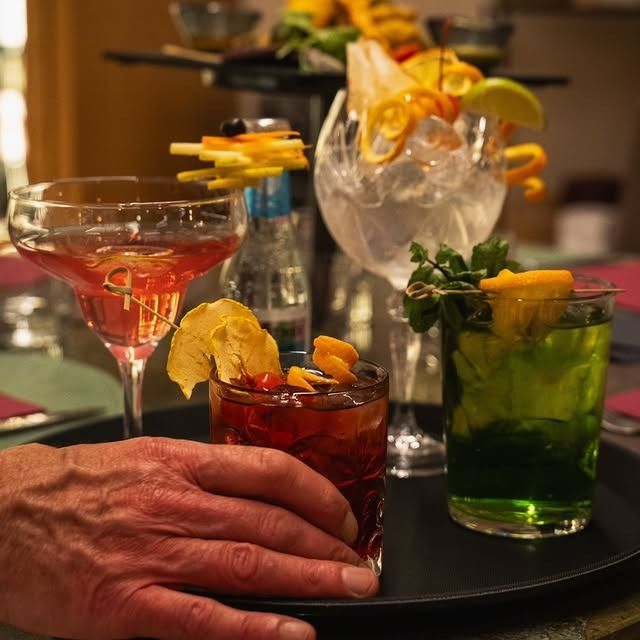 Mano di una persona che regge un vassoio con quattro cocktail colorati, guarniti con frutta, in un ambiente scarsamente illuminato.