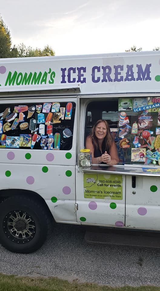 A smiling person leans out of the service window of a white ice cream truck decorated with green and purple polka dots.