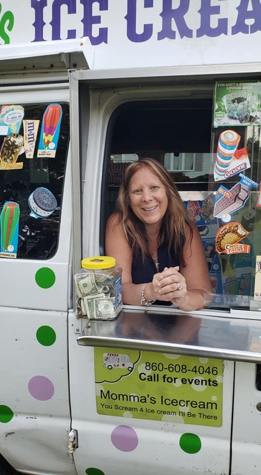 A smiling person leans out the window of a white Momma’s Ice Cream truck with a tip jar on the service counter.