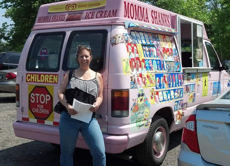 A woman stands smiling in front of a pink ice cream van decorated with colorful product signs in a parking lot.