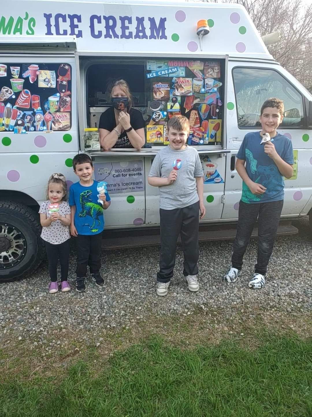 A person standing in an ice cream truck window smiles behind four children holding ice cream treats outdoors.