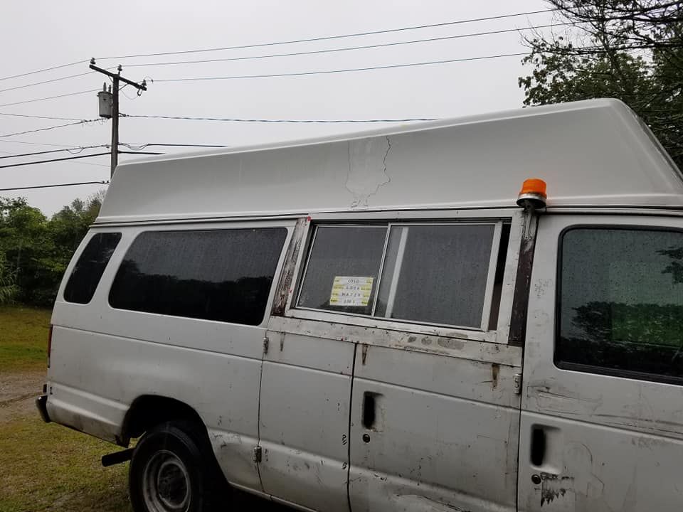 A white utility van with a raised roof and a small orange strobe light on its side, parked outdoors on a grassy surface.