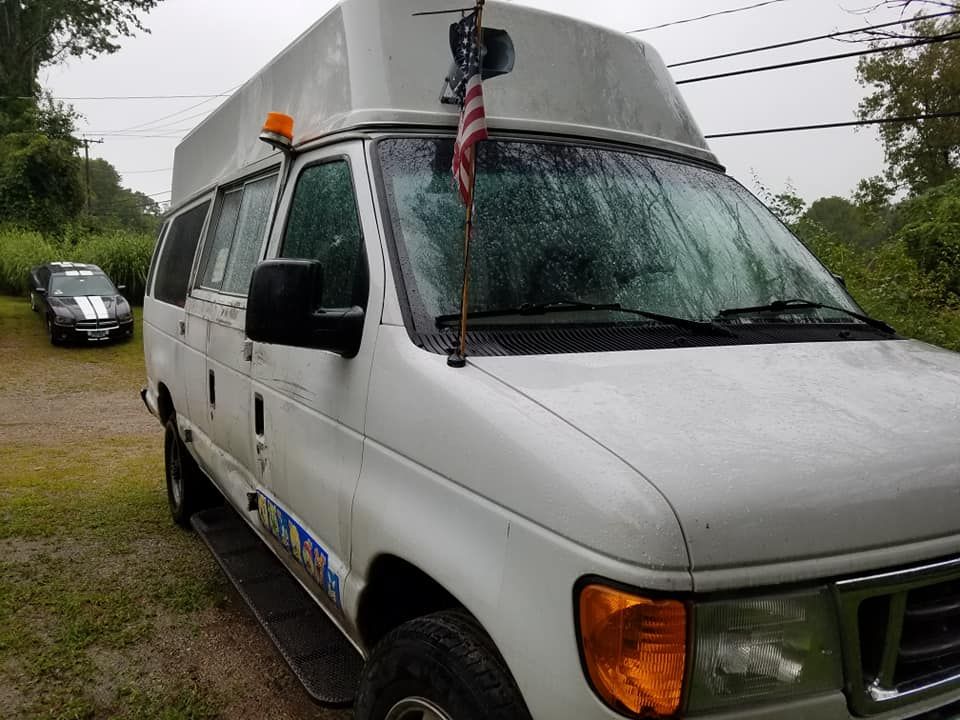 A white Ford van with a high roof, an orange light, and an American flag parked outdoors on a gravel lot.