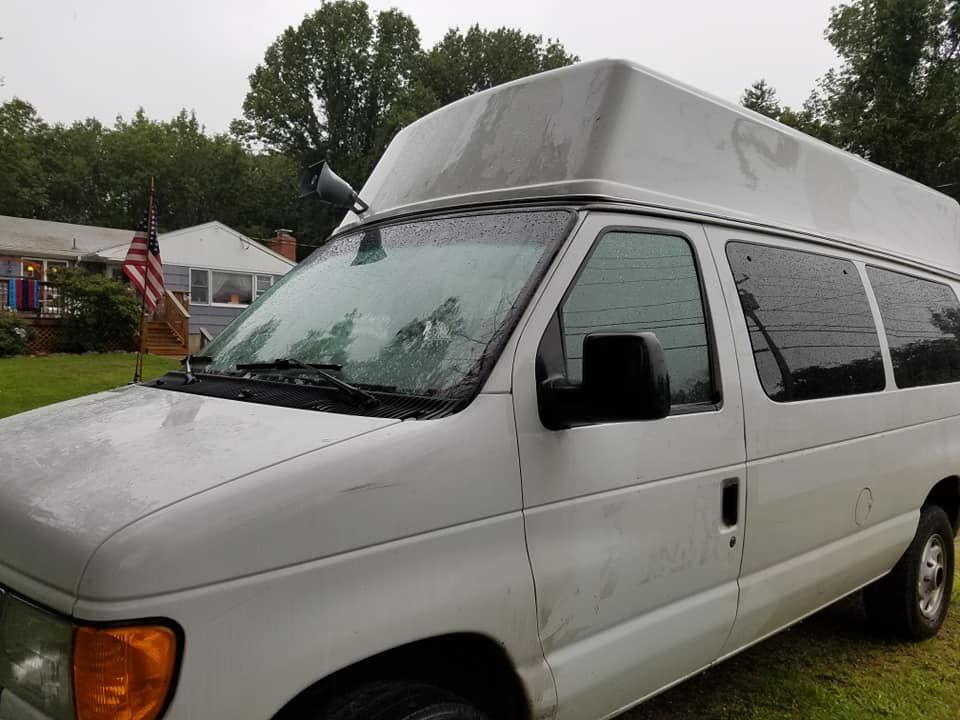 A white high-top Ford E-Series van parked on a grassy lawn with an American flag visible in the background.