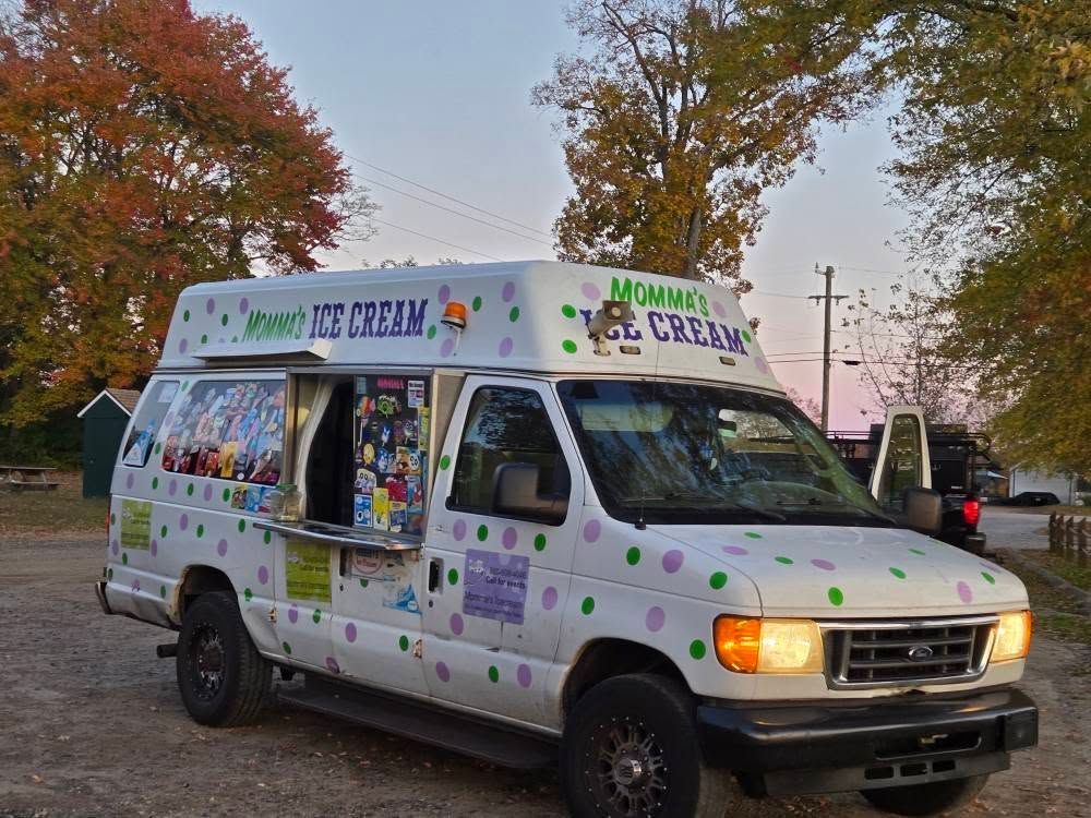 A white ice cream truck with purple and green polka dots parked outdoors with trees in the background.