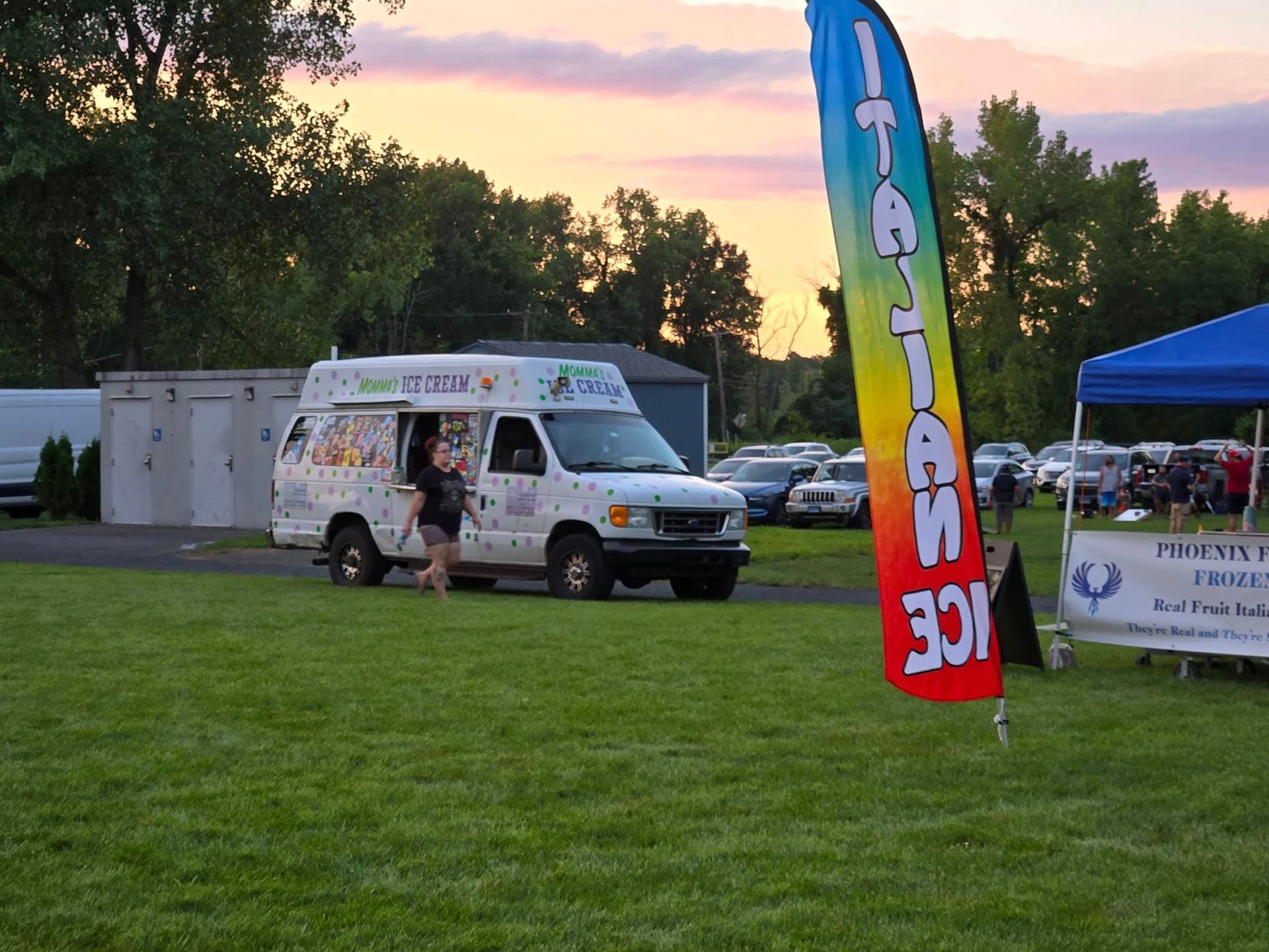 An ice cream truck parked on a grass field at sunset, next to a large red, yellow, and blue Italian ice promotional flag.