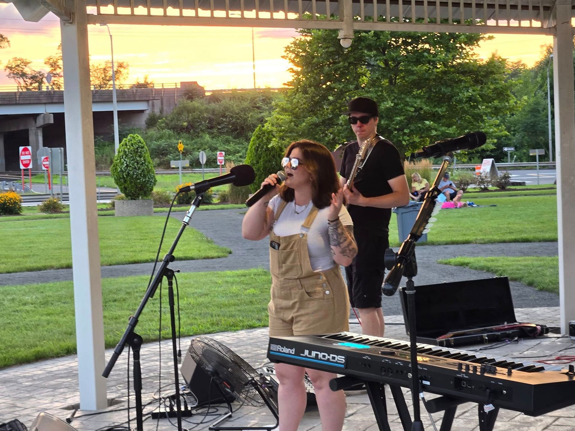 A person singing into a microphone next to a guitarist and keyboard at an outdoor gazebo concert during sunset.