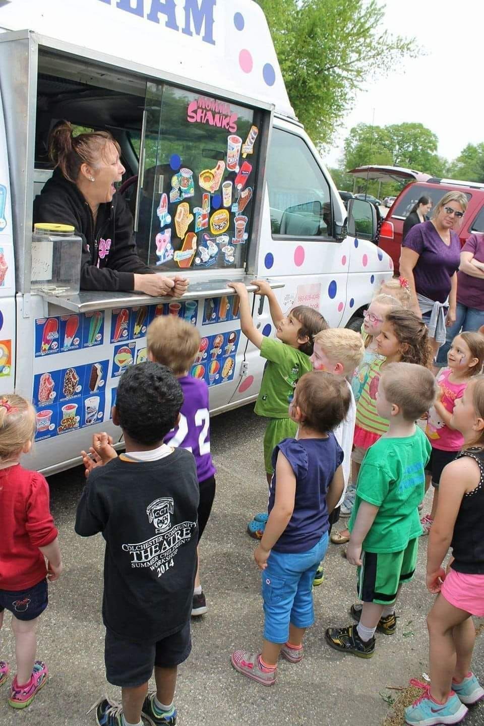 A smiling woman serves ice cream from a white food truck to a group of children waiting in line outdoors.