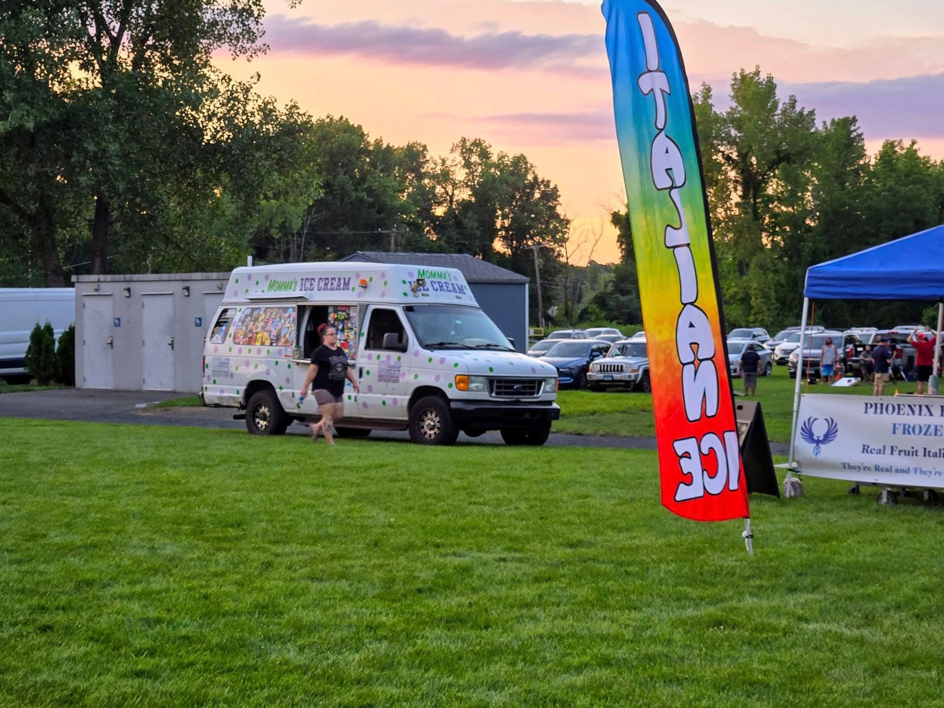 An ice cream truck drives on a grassy field at sunset near a large Italian Ice flag and a blue tent.