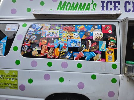 A white ice cream truck with purple and green polka dots features a menu window filled with colorful character popsicles.
