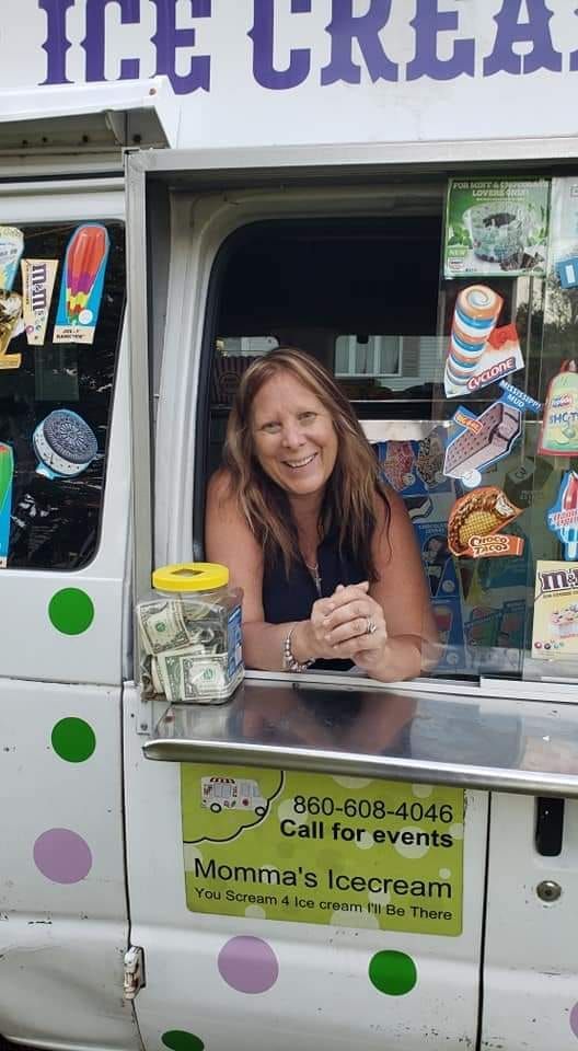 A smiling woman stands at the serving window of a white ice cream truck with a tip jar on the counter.