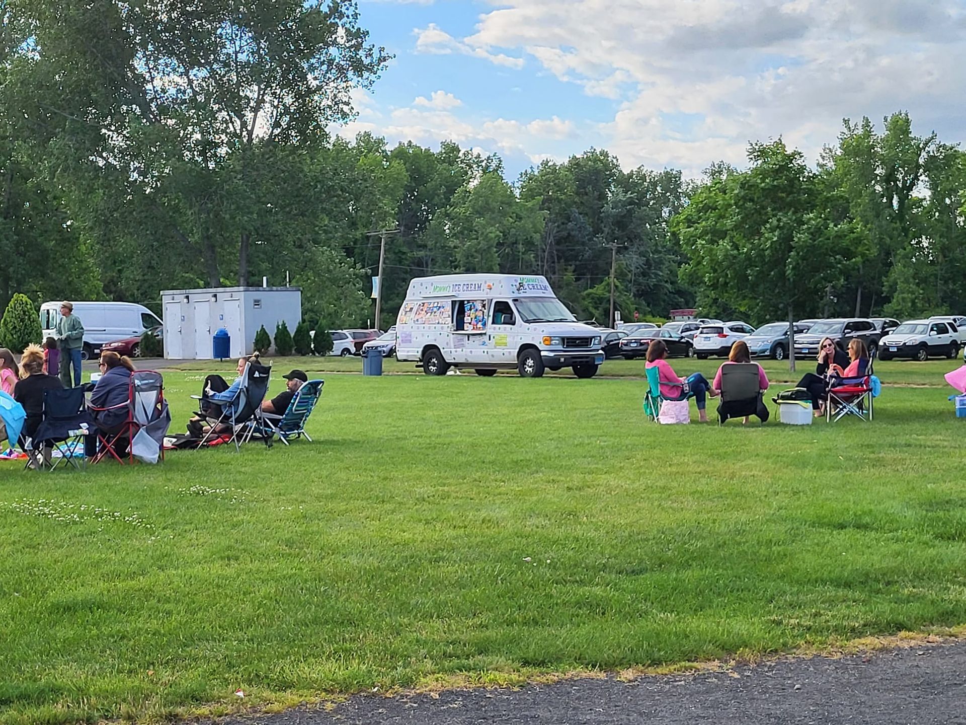 People sit in folding chairs on a green lawn near an ice cream truck parked in a gravel lot on a sunny day.