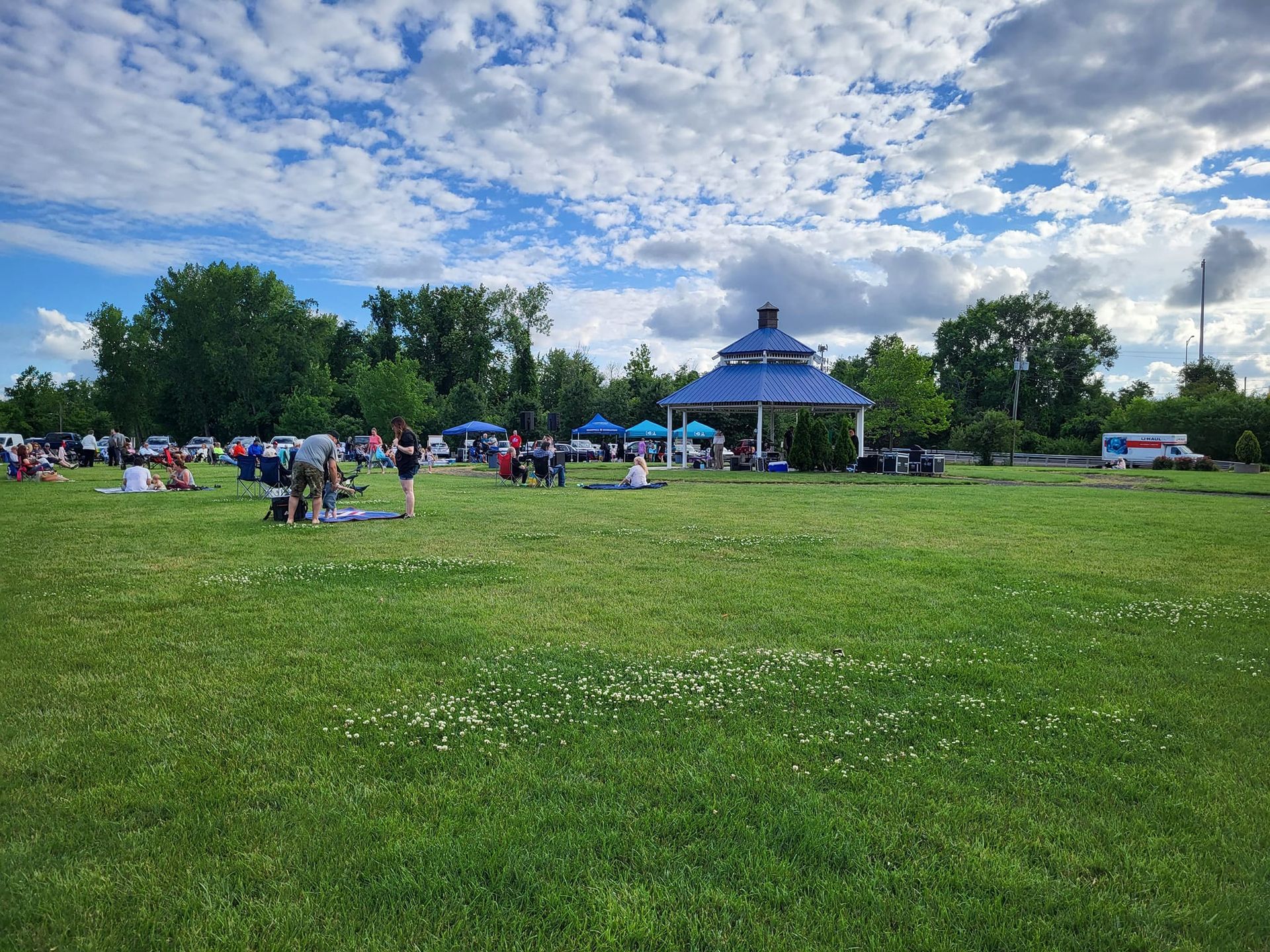People gather on a green park lawn in front of a blue-roofed gazebo under a sky with scattered clouds.