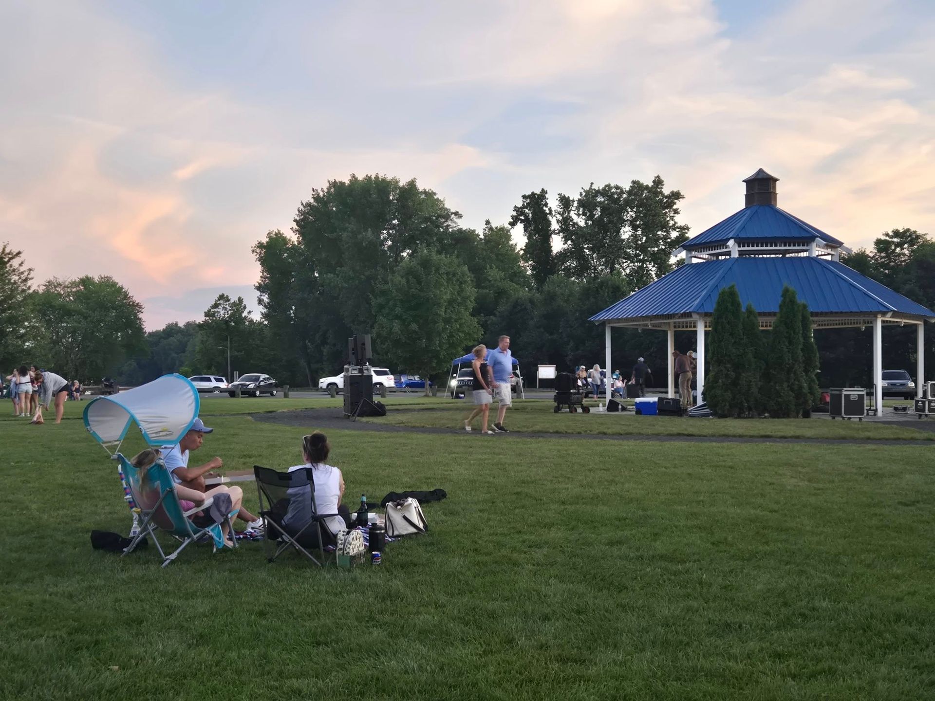People sit in chairs on a grassy lawn near a blue-roofed gazebo in a park during sunset.
