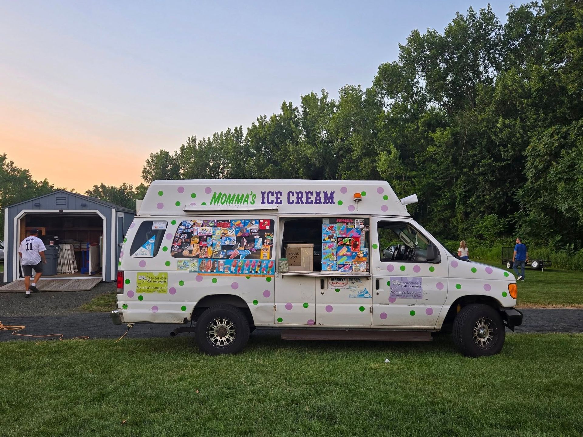 A white ice cream truck with colorful cartoon decals parked on a grassy field in front of a shed at sunset.