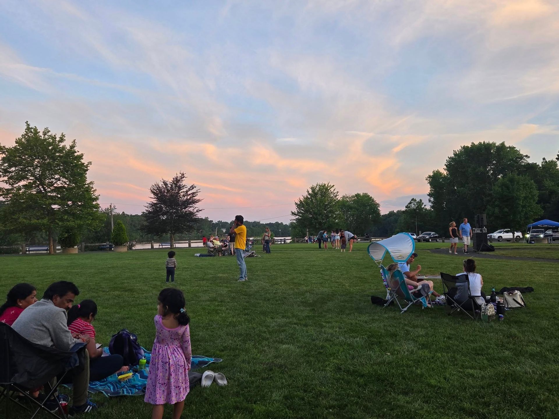 People relax and sit in folding chairs on a grassy park lawn under a vibrant, pastel sunset sky.