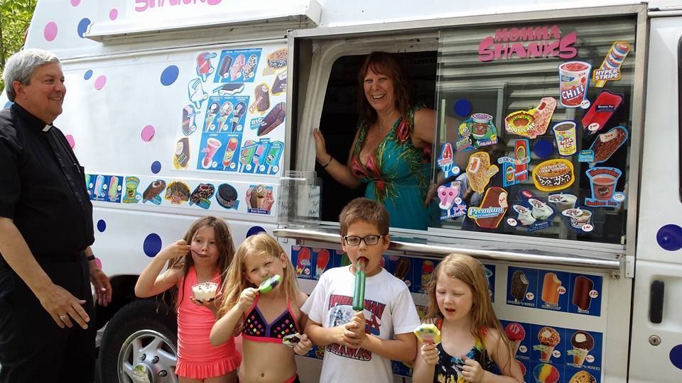 A woman serves treats from an ice cream truck to a group of children, while a man in a black clerical shirt stands by.