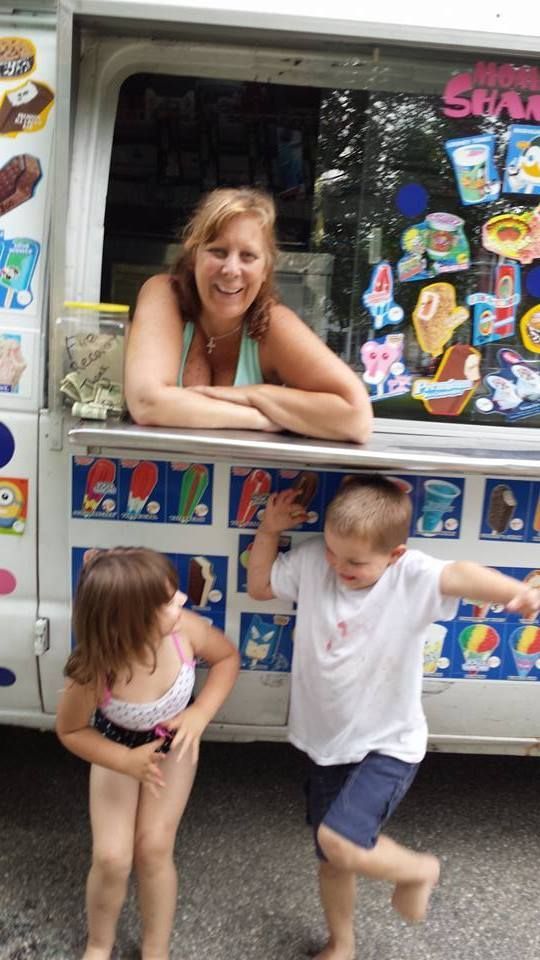 A smiling person leans out of an ice cream truck window as a child in a swimsuit and a child in a white shirt pose nearby.