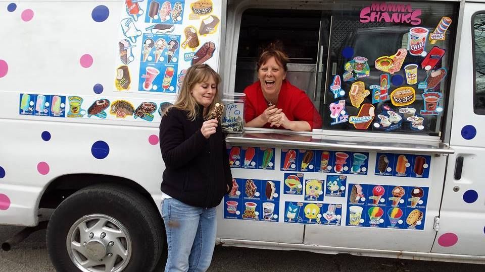 A person eats an ice cream cone outside an ice cream truck, while an employee smiles from the service window.
