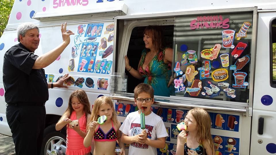 A man waves in front of an ice cream truck while a woman serves three children eating popsicles at the service window.