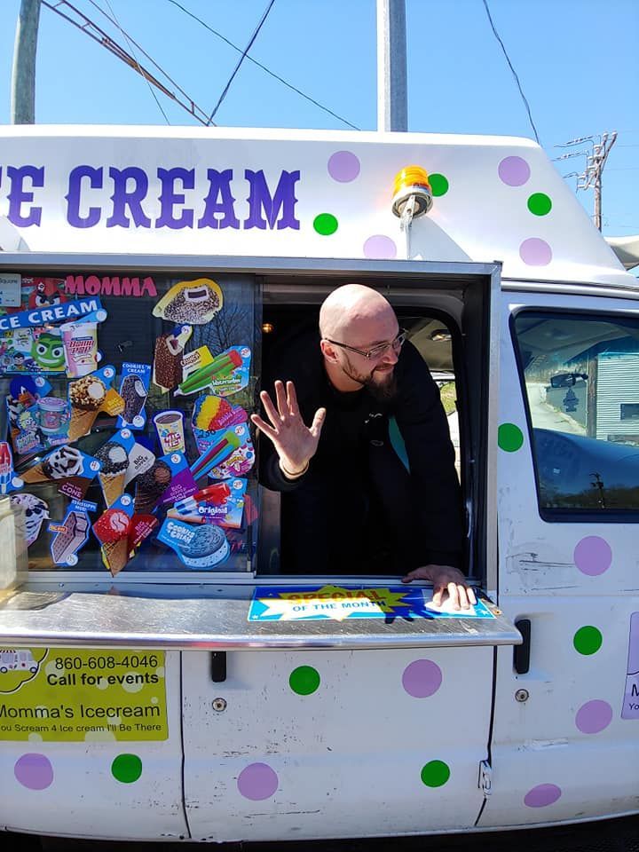 A person with a beard and glasses waves from the window of a white ice cream truck decorated with colorful polka dots.